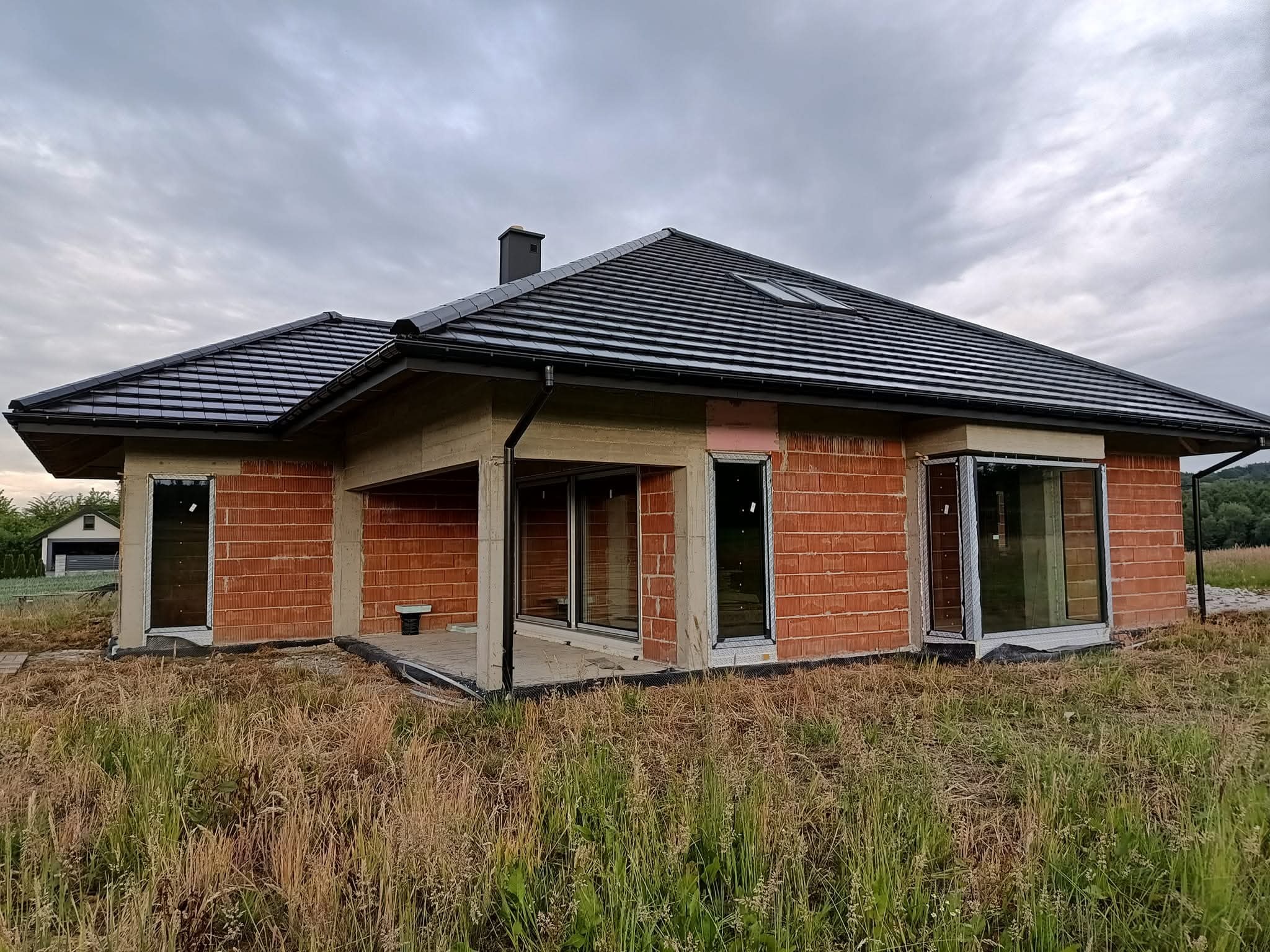Dom jednorodzinny w stanie surowym otwartym z czarną dachówką i widocznymi oknami oraz drzwiami. Niski kąt ujęcia, pochmurne niebo.