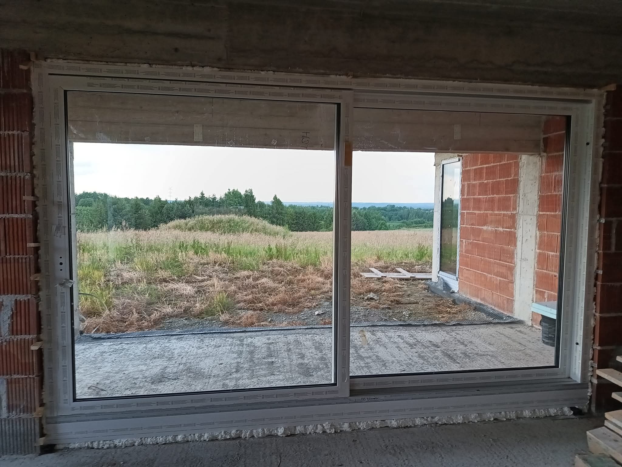 Białe okno przesuwne PCV zamontowane w surowym murze z widokiem na łąkę i las. Widoczne ślady pianki montażowej i niedokończone wykończenie.