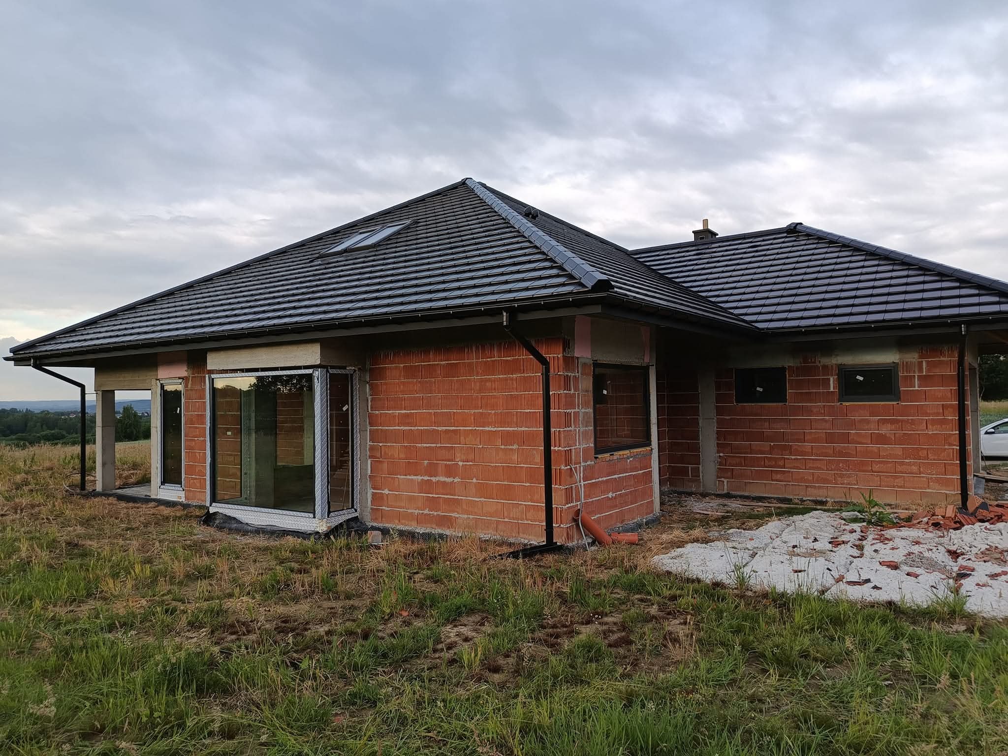 Dom w trakcie budowy z widocznymi oknami PCV, murowanymi ścianami i ciemną dachówką. Widok na zielony teren i pochmurne niebo w tle.