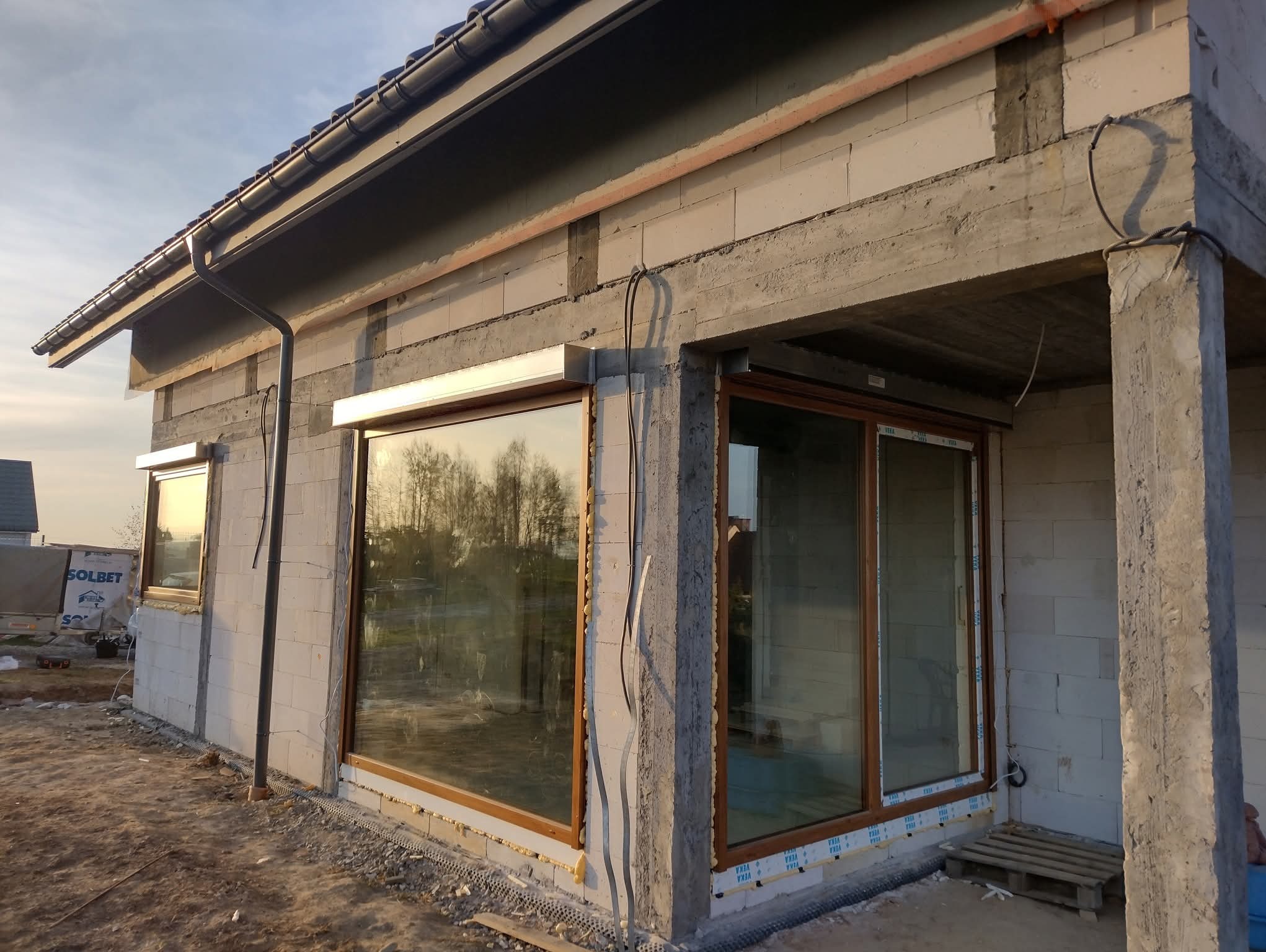 Dom w trakcie budowy z zamontowanymi oknami z roletami zewnętrznymi i widoczną konstrukcją z betonu komórkowego. Widoczne okna z brązowymi ramami.