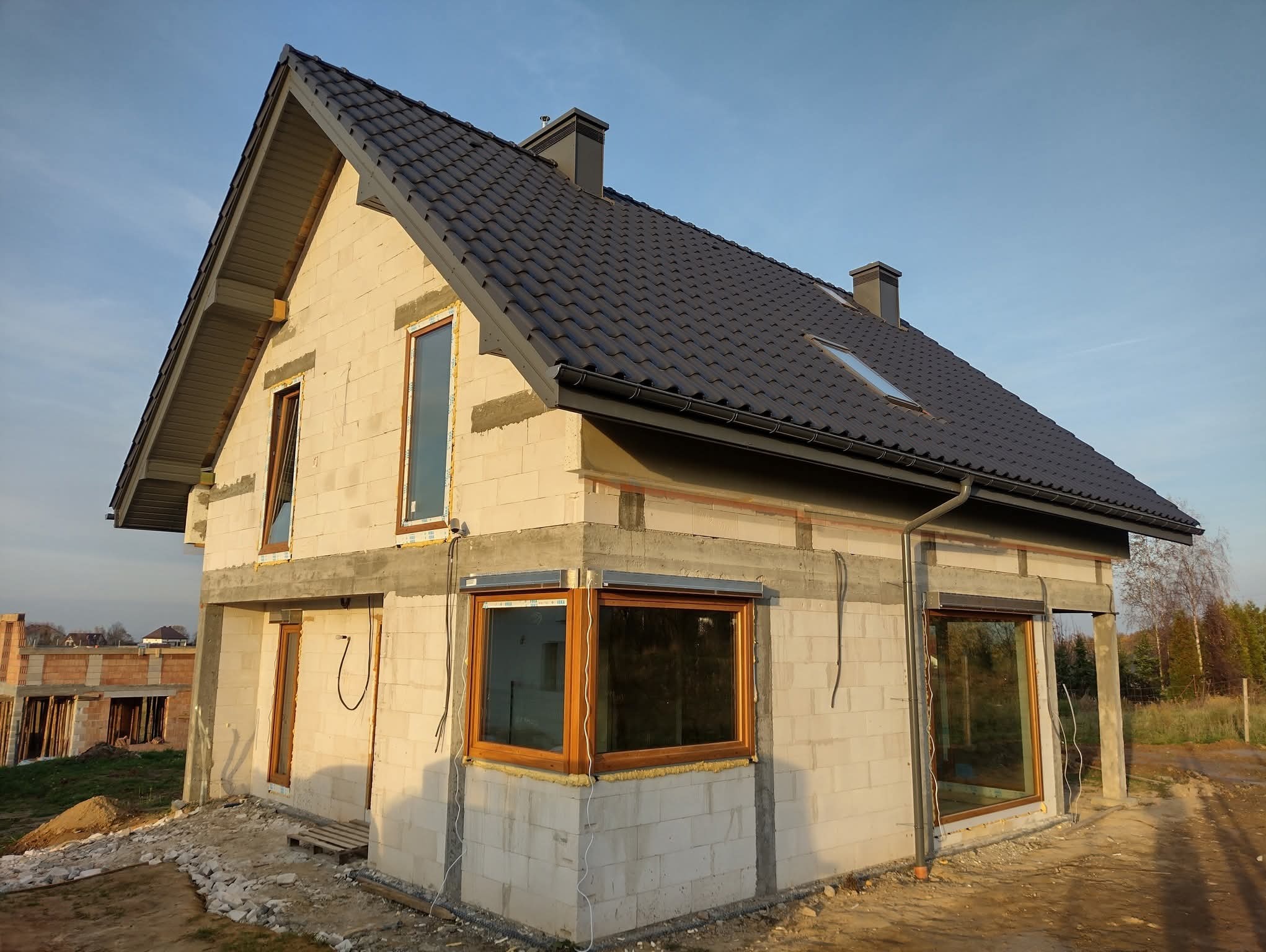 Dom w trakcie budowy z zamontowanymi oknami z drewnianą ramą i roletami zewnętrznymi. Widoczny surowy stan ścian i dachu z blachy trapezowej.