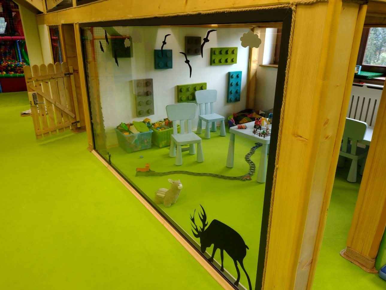 Przeszklona konstrukcja z drewnianą ramą, imitująca pokój zabaw dla dzieci z zabawkami, naklejkami zwierząt i klockami na ścianie. Widoczne elementy placu zabaw.