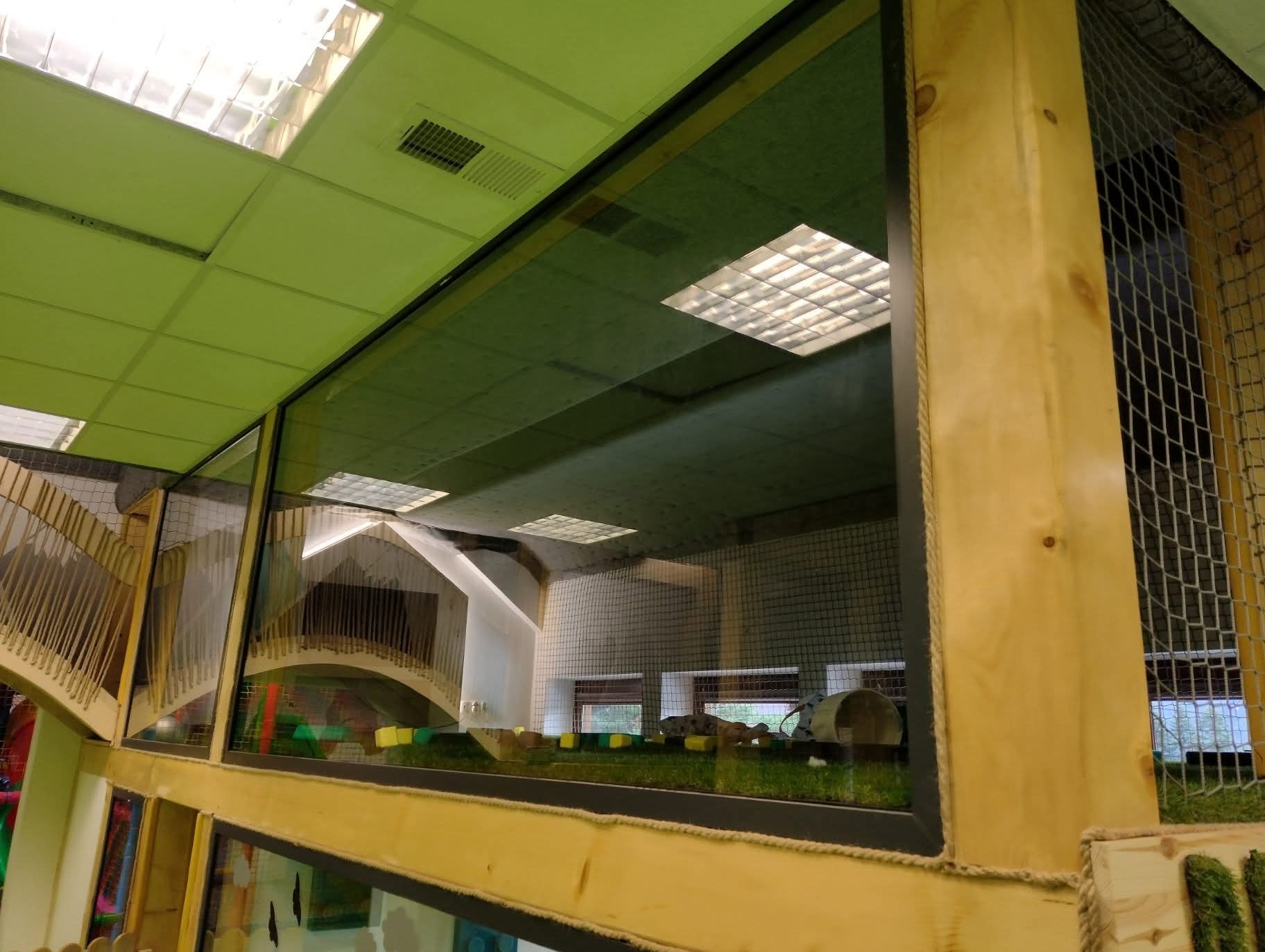Szklana przegroda z drewnianą ramą, oddzielająca przestrzeń zabaw dla dzieci. Widoczne schody, mostki i sztuczna trawa wewnątrz. Oświetlenie sufitowe.