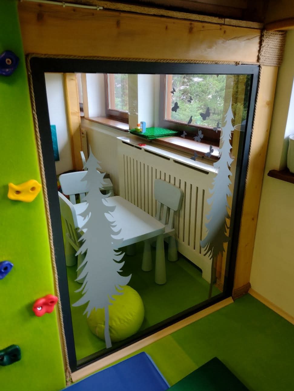 Kreatywnie zaaranżowane okno z naklejkami drzew i motyli, widok na pokój dziecięcy z zieloną ścianą do wspinaczki i meblami w jasnych kolorach.