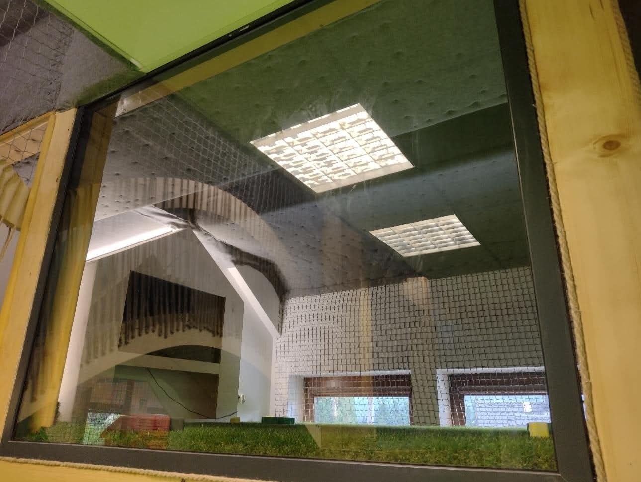 Czarna rama okienna z widokiem na wnętrze budynku z siatką ochronną. W odbiciu widoczna klatka schodowa i oświetlenie sufitowe.