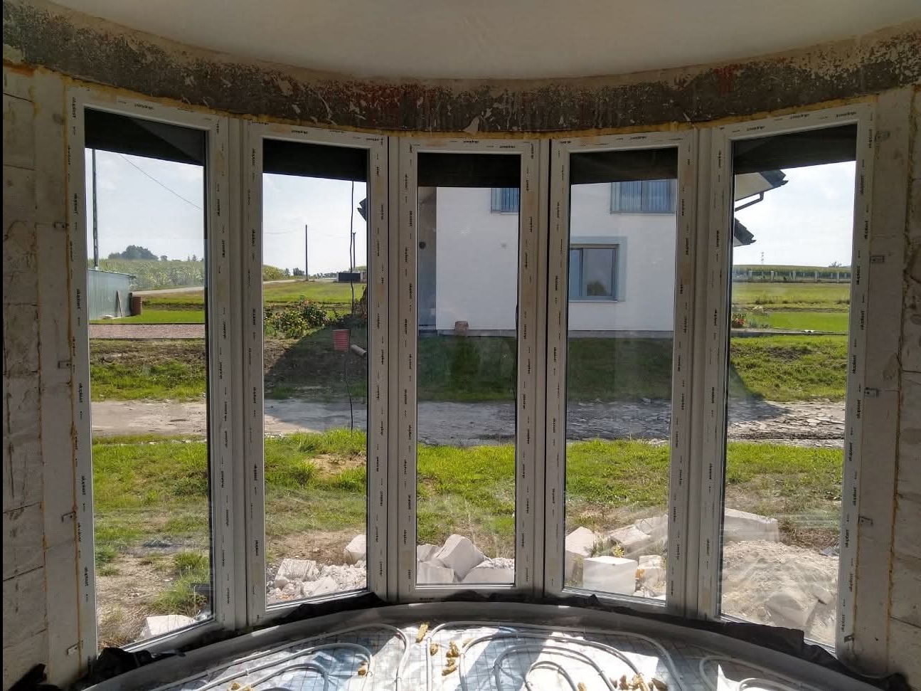 Wnętrze w trakcie wykańczania z zamontowanymi białymi oknami PCV, widok na zewnątrz na dom i zieleń. Elementy instalacji ogrzewania podłogowego w dolnej części kadru.