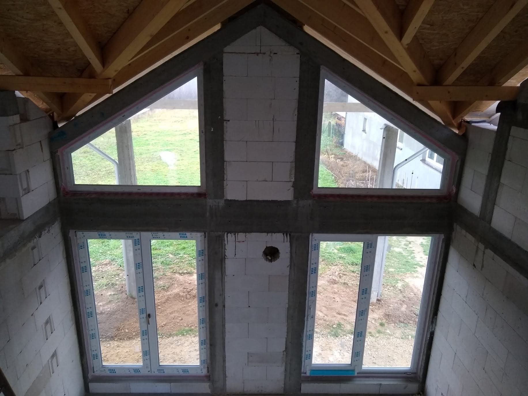 Montaż okien PCV w surowym stanie domu z widoczną konstrukcją dachu i bloczkami. Dwa okna trójkątne na górze i dwa prostokątne na dole. Widok na zewnątrz.