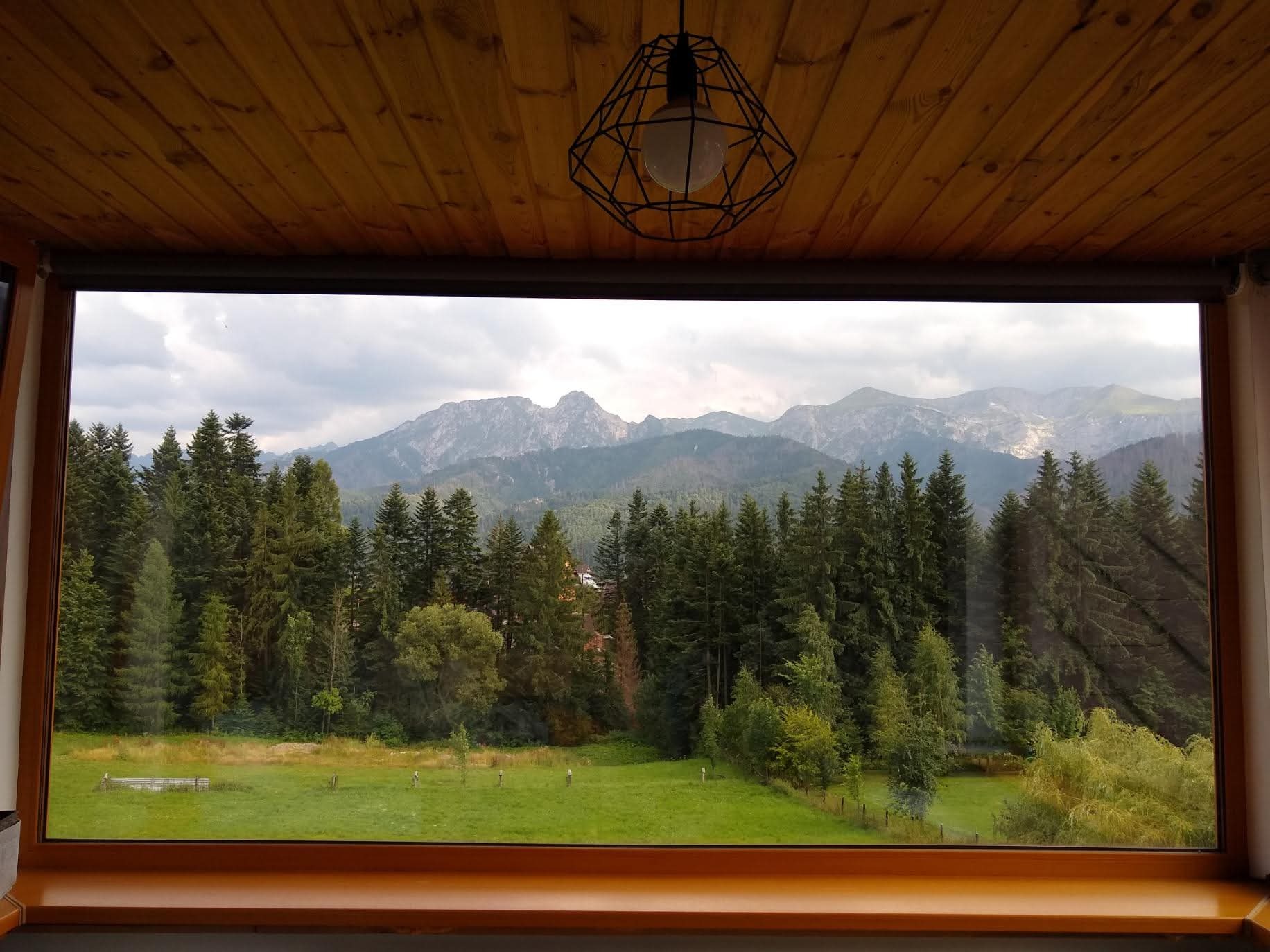 Widok Tatr przez duże okno z drewnianą ramą i żyrandolem. Panorama gór, las i łąka za oknem. Drewniany sufit nad oknem.