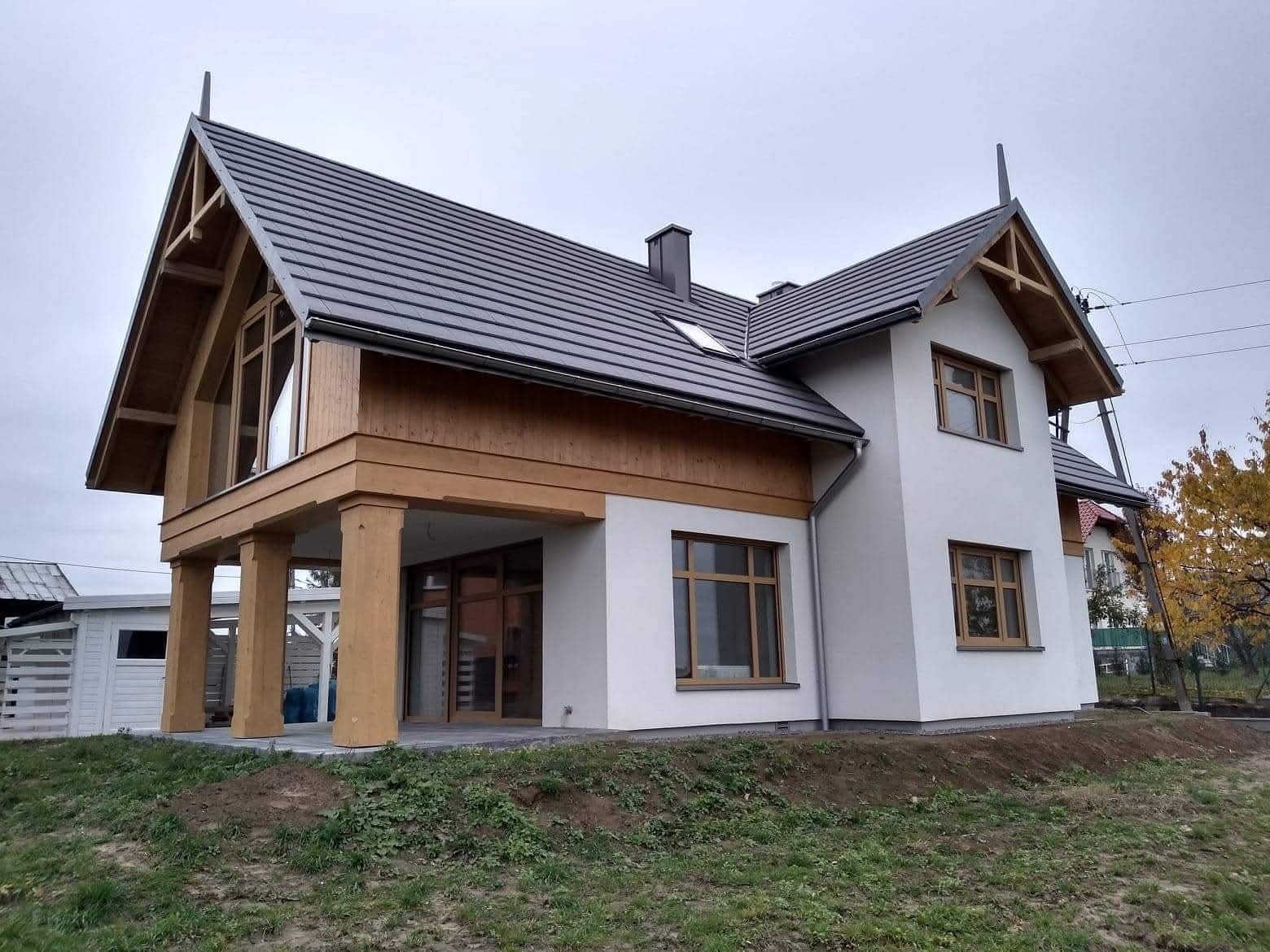 Nowoczesny dom jednorodzinny z grafitową dachówką i drewnianymi elementami wykończeniowymi w Brzesku. Widok z zewnątrz, jesienna sceneria.