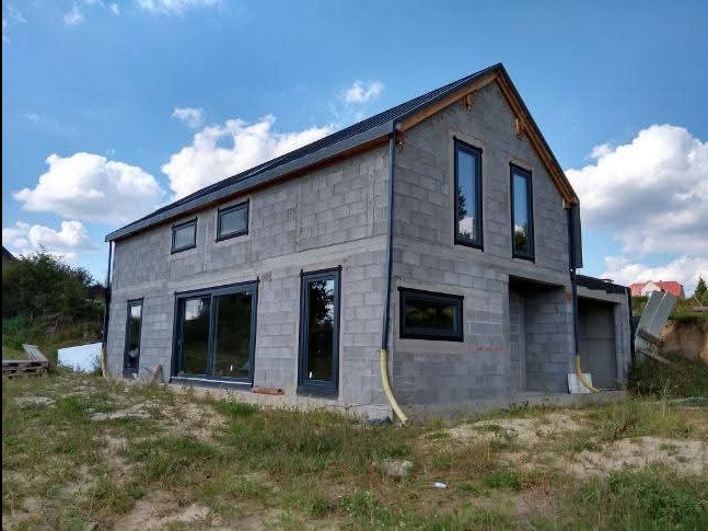 Surowy dom z szarego betonu komórkowego z czarnymi oknami, w trakcie budowy, na tle błękitnego nieba z chmurami. Widoczny fragment dachu i otoczenie placu budowy.