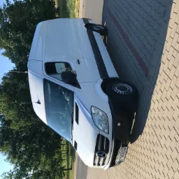 Biały samochód dostawczy Mercedes-Benz Sprinter zaparkowany na brukowanej powierzchni w słoneczny dzień, widok z przodu na tle drzew.