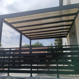 Spawmar - Nowoczesna pergola z przezroczystym zadaszeniem, widok zza czarnego, poziomego ogrodzenia, w tle drzewa i błękitne niebo.