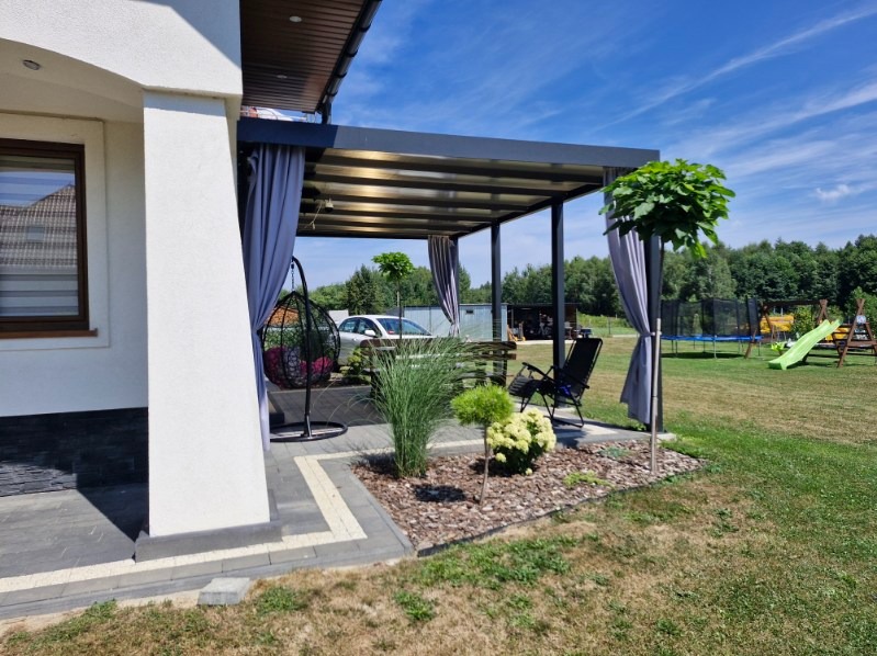 Zadaszona pergola aluminiowa z szarymi zasłonami, wiszącym fotelem i starannie zagospodarowanym ogrodem, widok na dom z oknem i plac zabaw w tle.