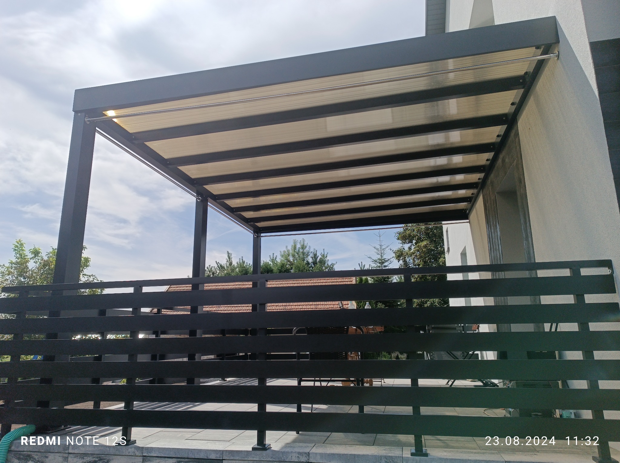 Nowoczesna pergola z przezroczystym zadaszeniem, widok zza czarnego, poziomego ogrodzenia, w tle drzewa i błękitne niebo.