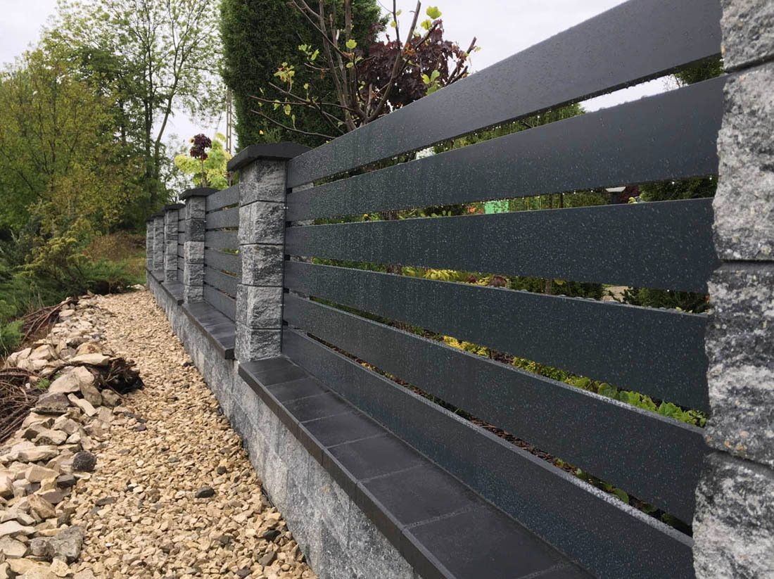 Nowoczesne ogrodzenie panelowe z poziomych, grafitowych desek, wsparte na kamiennych słupkach, z widocznymi kroplami deszczu na powierzchni, otoczone żwirowym podłożem i zielenią.