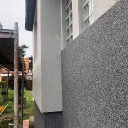 Fragment elewacji domu z oknami z luksferów, pokrytej tynkiem mozaikowym w odcieniach szarości, widoczne rusztowanie.