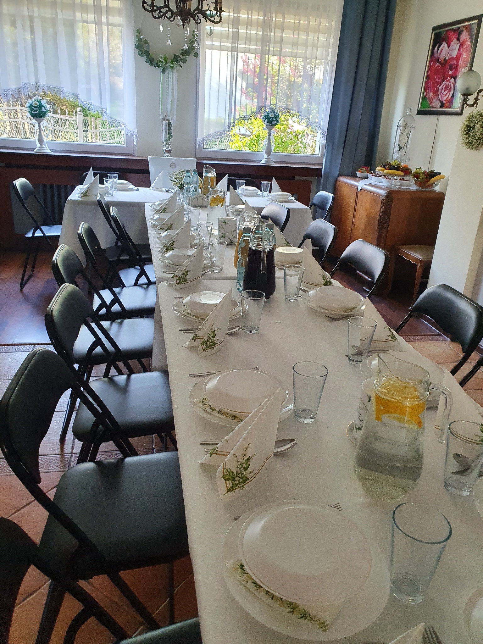 Uroczysty stół z białym obrusem, nakryty dla wielu gości, z dekoracjami kwiatowymi na serwetkach i dzbankami z napojami, ustawiony w jasnym pomieszczeniu z oknami.