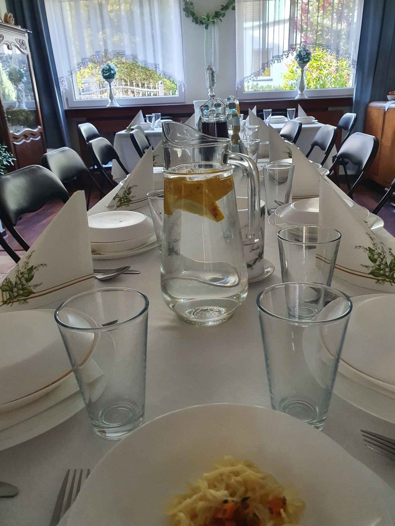 Ustawiony stół z białym obrusem, nakryciami, karafką z wodą i cytryną, oraz dekoracyjnymi serwetkami z motywem konwalii, w tle okno z białymi firankami i dekoracjami roślinnymi.