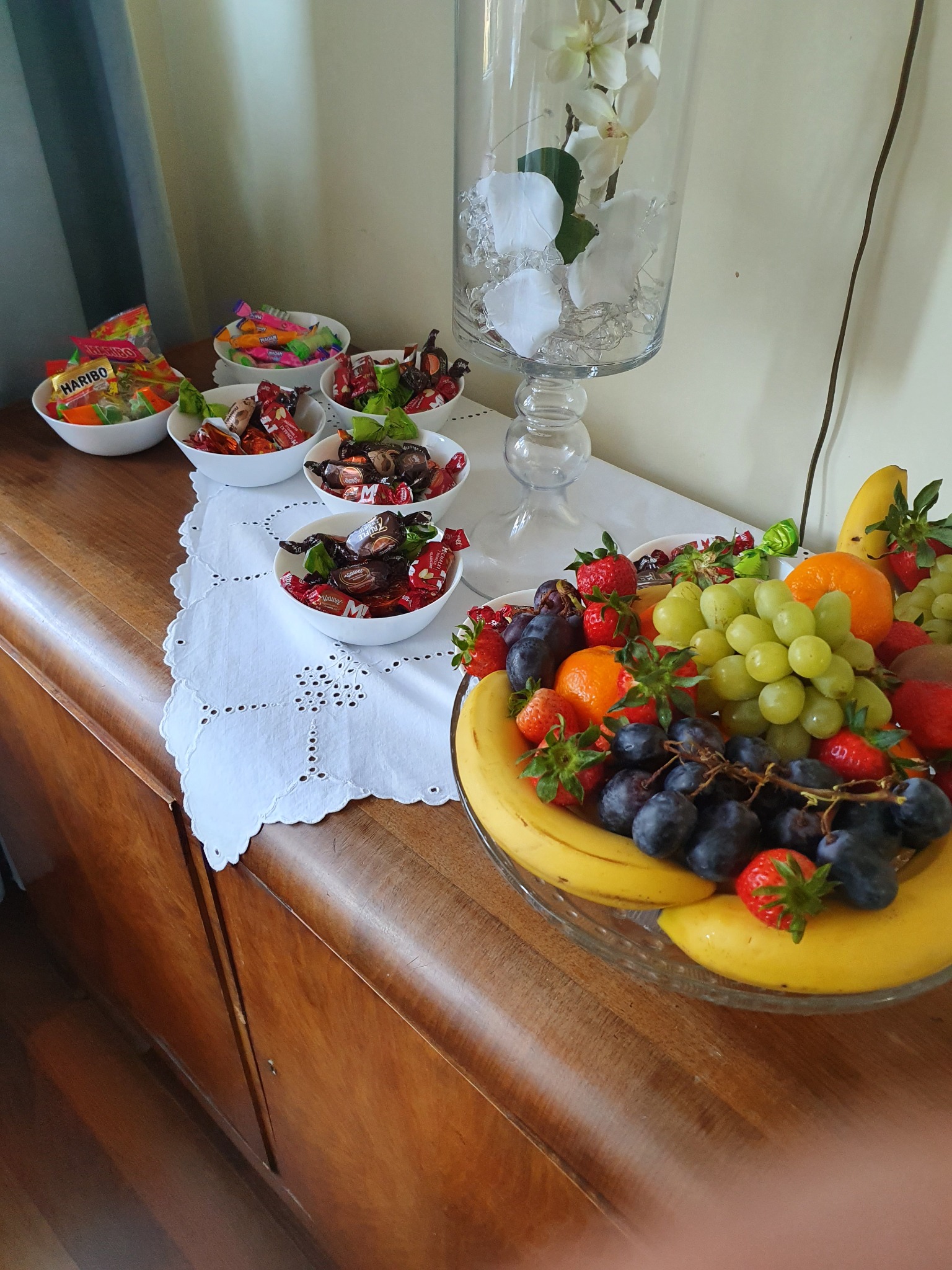 Stół z drewnianymi szafkami, na którym znajdują się miseczki z cukierkami i szklana misa z bananami, winogronami, truskawkami i pomarańczami oraz wazon z kwiatami i ozdobnymi kamykami.