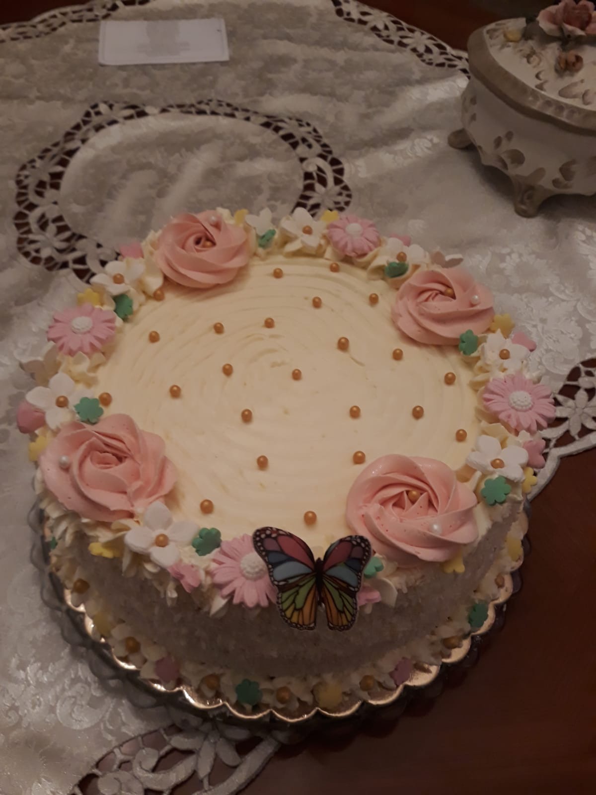 Dekorowany tort z kremem, różowymi różami, małymi kwiatkami i motylem, na stole z koronkową serwetą