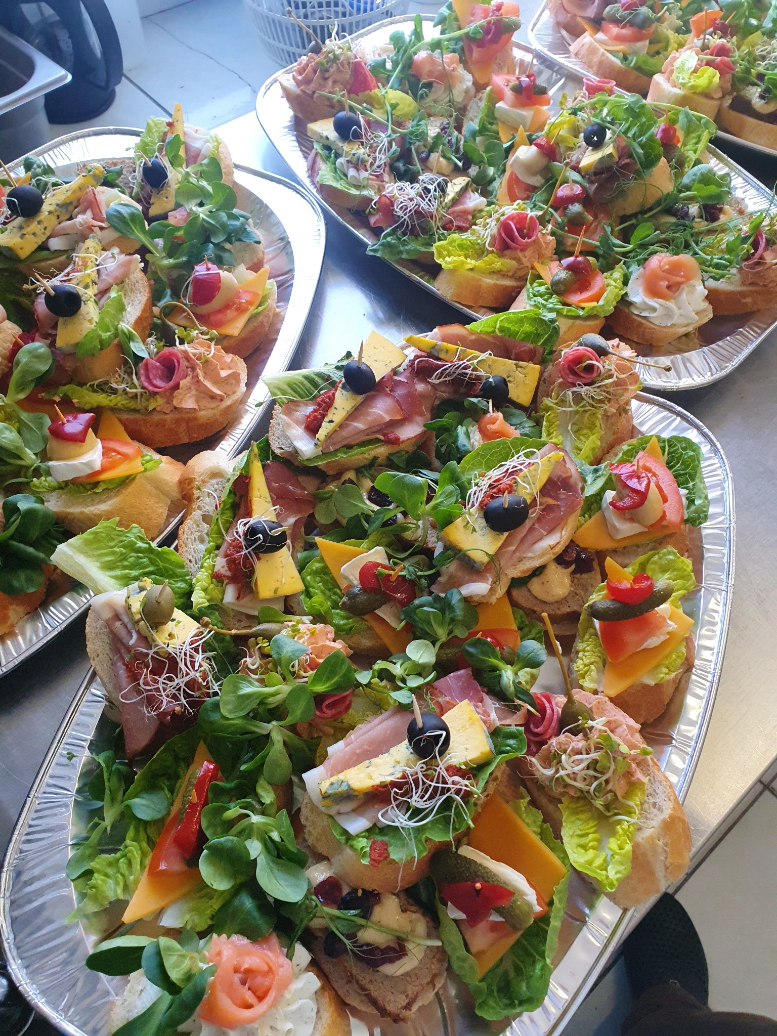 Srebrne tace pełne różnorodnych kanapek z wędlinami, serami, warzywami i ziołami, ułożone artystycznie na stole.