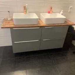 Nowoczesna łazienka z dwoma umywalkami nablatowymi o kwadratowym kształcie i miedzianymi bateriami, szara szafka z czterema szufladami, ciemna podłoga i białe płytki na ścianie z delikatnym wzorem...