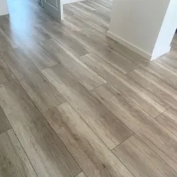 Ułożona podłoga z paneli imitujących drewno w odcieniach szarości, widoczny fragment białej ściany z listwą przypodłogową oraz przejście do innego pomieszczenia.