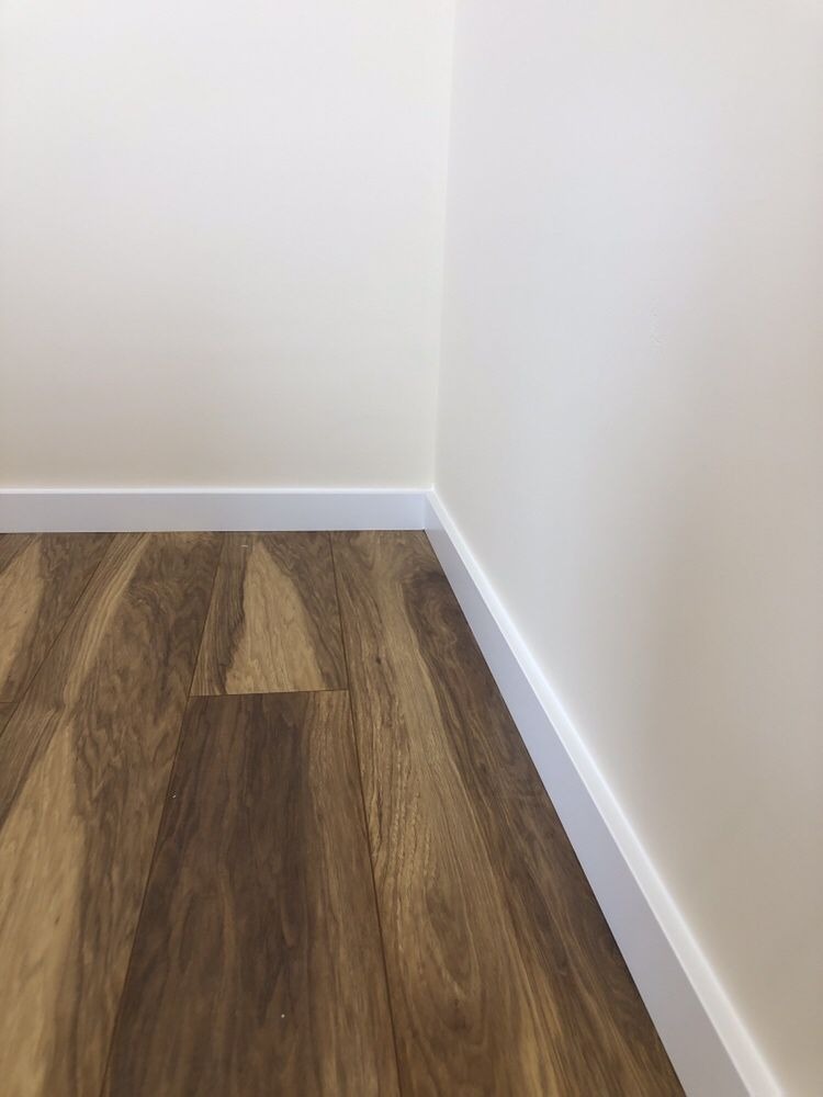 Ukończony róg pokoju z panelami podłogowymi imitującymi drewno i białą listwą przypodłogową przy jasnej ścianie.