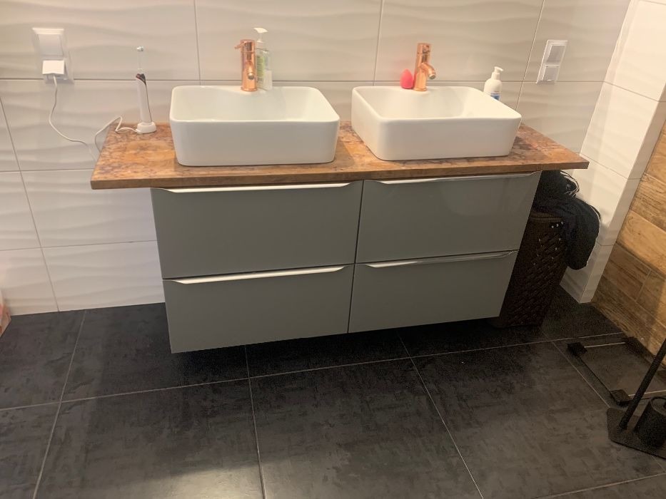 Nowoczesna łazienka z dwoma umywalkami nablatowymi o kwadratowym kształcie i miedzianymi bateriami, szara szafka z czterema szufladami, ciemna podłoga i białe płytki na ścianie z delikatnym wzorem...