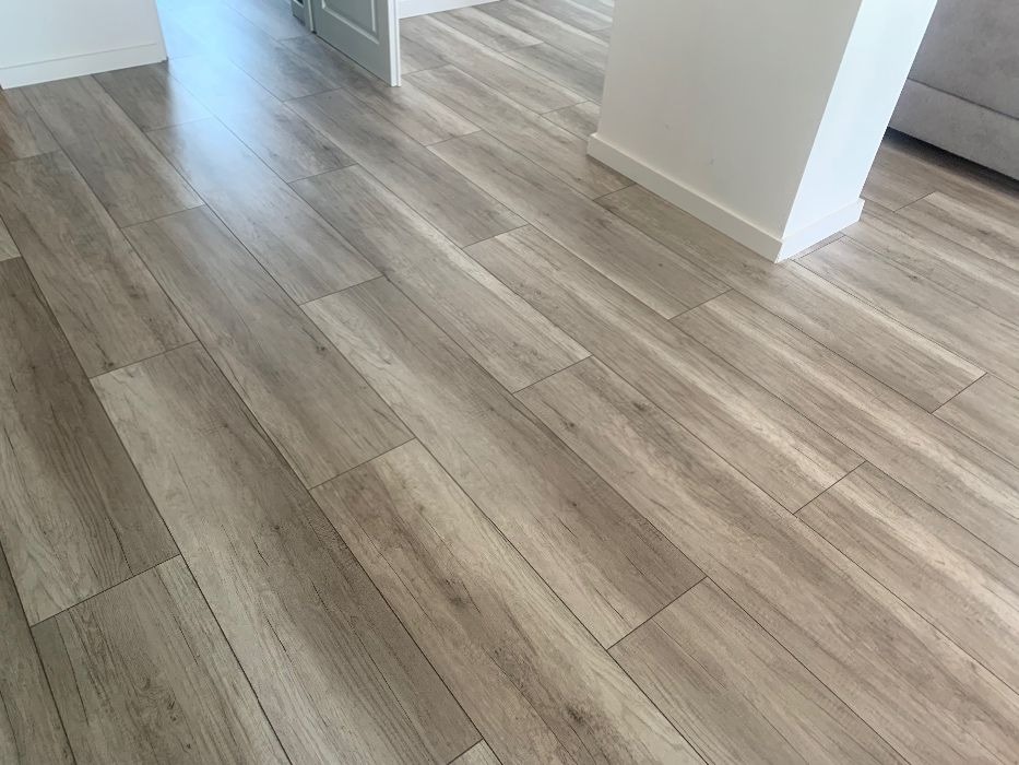 Ułożona podłoga z paneli imitujących drewno w odcieniach szarości, widoczny fragment białej ściany z listwą przypodłogową oraz przejście do innego pomieszczenia.