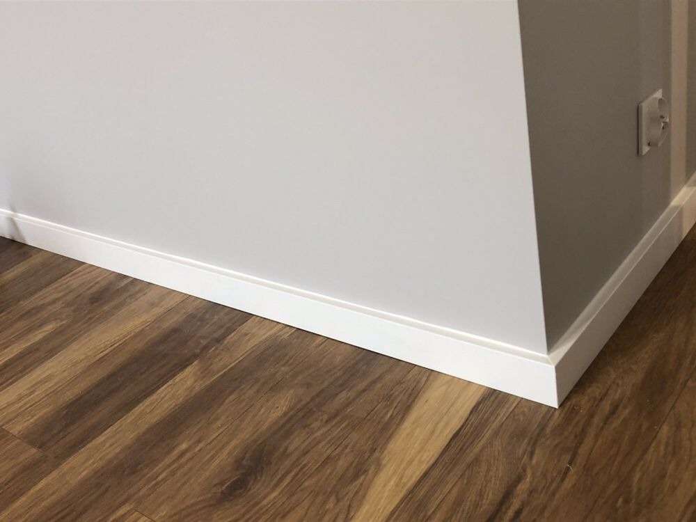 Wykończony narożnik pokoju z drewnopodobną podłogą, szarą ścianą i białą listwą przypodłogową, widoczne gniazdko elektryczne.