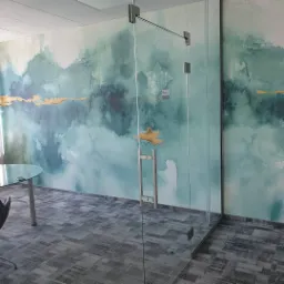 Szklana ścianka działowa w biurze z abstrakcyjnym wzorem w odcieniach błękitu i złota na ścianie, widoczny fragment okrągłego stołu i krzesła.