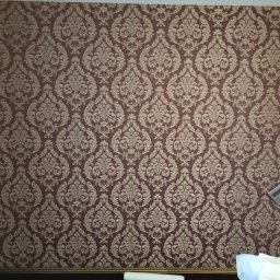 WŁW USŁUGI ŁUKASZ ZOWCZAK - Ściana pokryta bordową tapetą w złote, symetryczne wzory. Widoczne gniazdka elektryczne i fragmenty przedmiotów przy ścianie. Dekoracyjne wykończenie wnętrza.