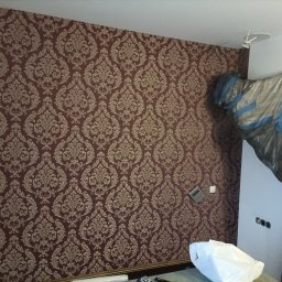 WŁW USŁUGI ŁUKASZ ZOWCZAK - Ściana po tapetowaniu w brązowo-złote wzory. Część pomieszczenia zabezpieczona folią malarską. Widoczne gniazdka elektryczne i wystający element oświetlenia sufitowego.