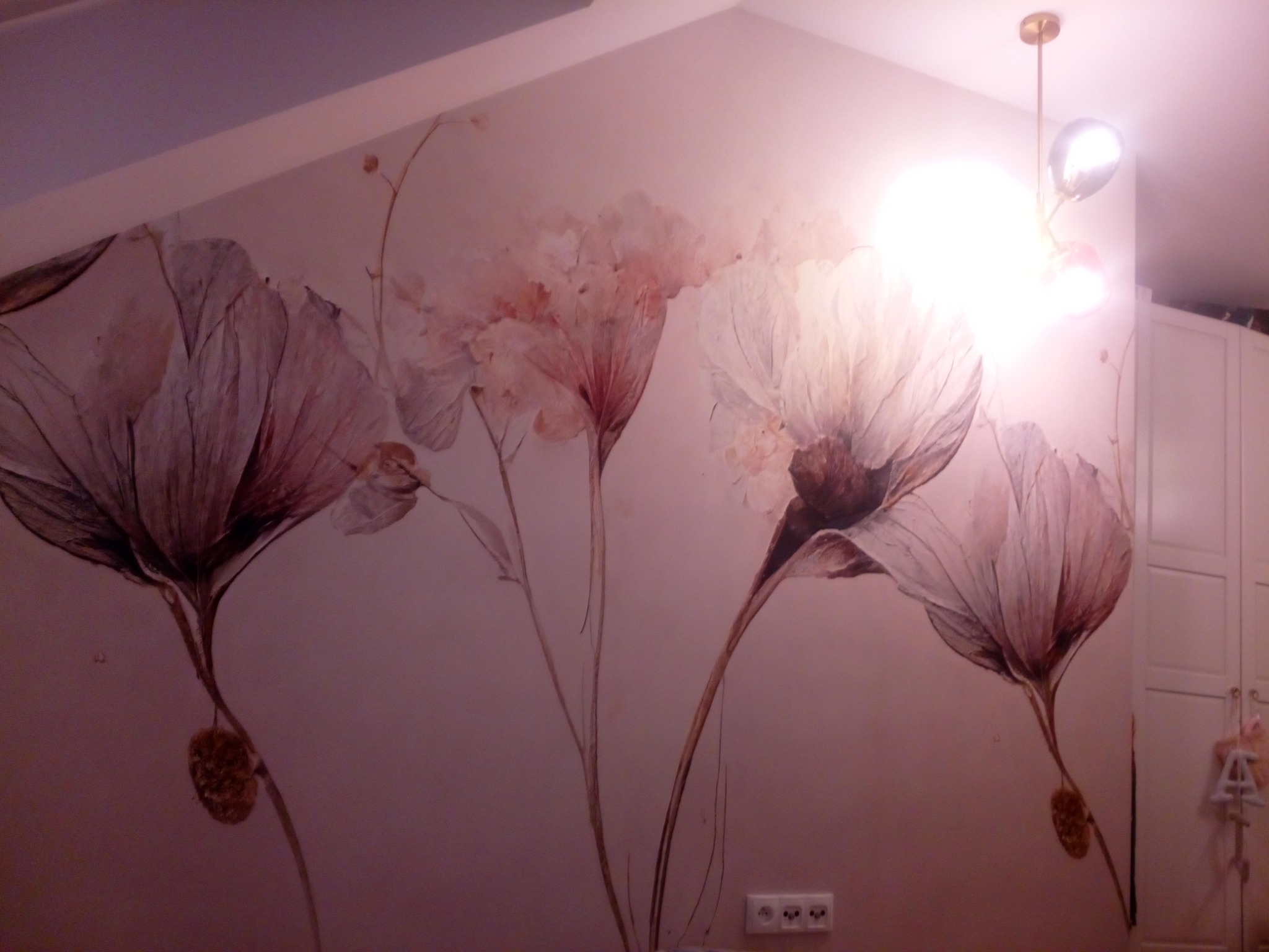 Ściana w pokoju z fototapetą przedstawiającą duże, stylizowane kwiaty w odcieniach różu i brązu, oświetlona przez lampę sufitową z trzema kloszami. Widoczny fragment gniazdka elektrycznego przy...