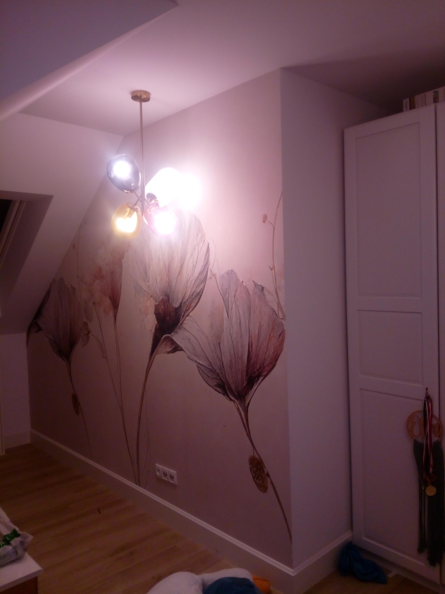 Ściana w pokoju na poddaszu z fototapetą przedstawiającą kwiaty, oświetlona nowoczesną lampą wiszącą z kulistymi kloszami, obok biała szafa.