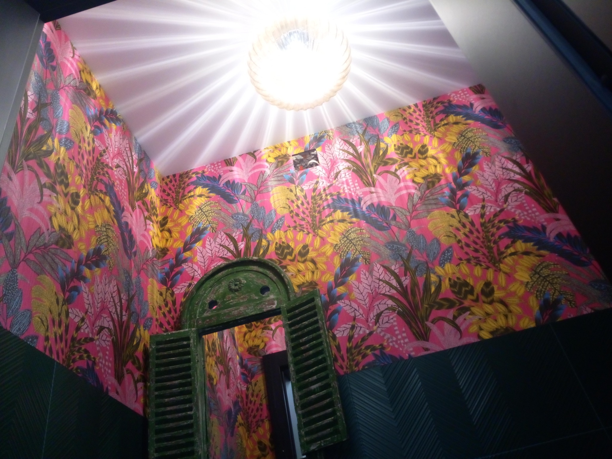 Wnętrze łazienki z intensywną, tropikalną tapetą w odcieniach różu, żółci i zieleni, uzupełnione zieloną, drewnianą ramą okienną z okiennicami i ciemnozielonymi płytkami u dołu ściany. Oświetlenie...