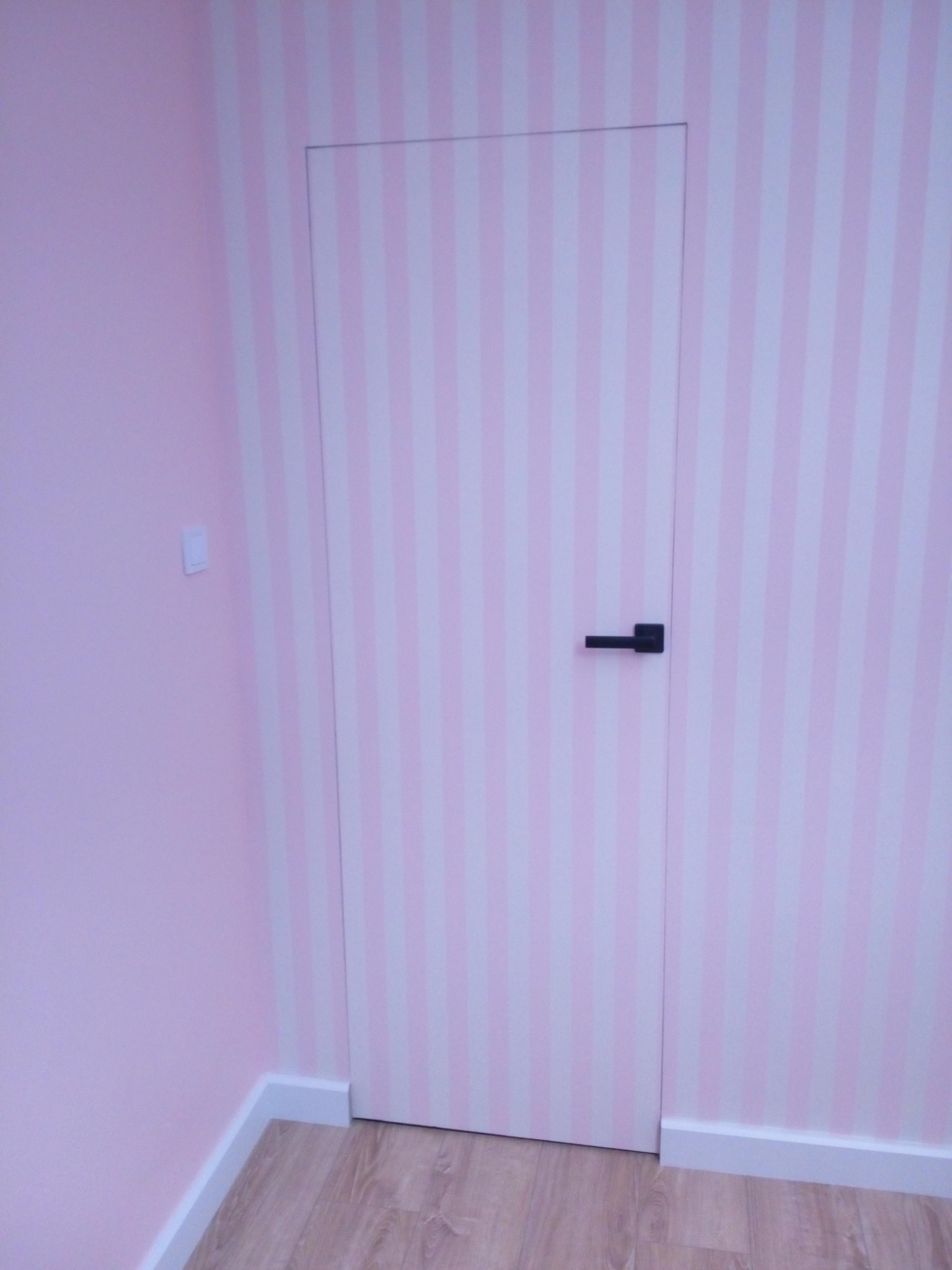 Różowo-białe pionowe pasy na ścianie i ukrytych drzwiach z czarną klamką, dopasowane do koloru ścian, listwa przypodłogowa w kolorze białym, jasna drewniana podłoga.