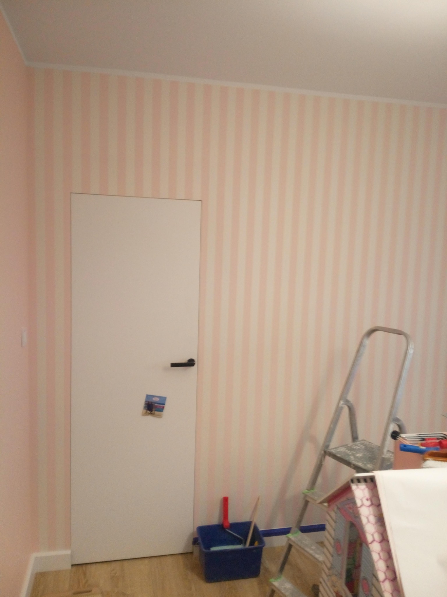 Pokój w trakcie odświeżania: ściany w pastelowe pionowe pasy, drabina oparta o ścianę, obok wiadro z farbą i wałkiem, częściowo zasłonięte kartonową konstrukcją domku dla lalek.