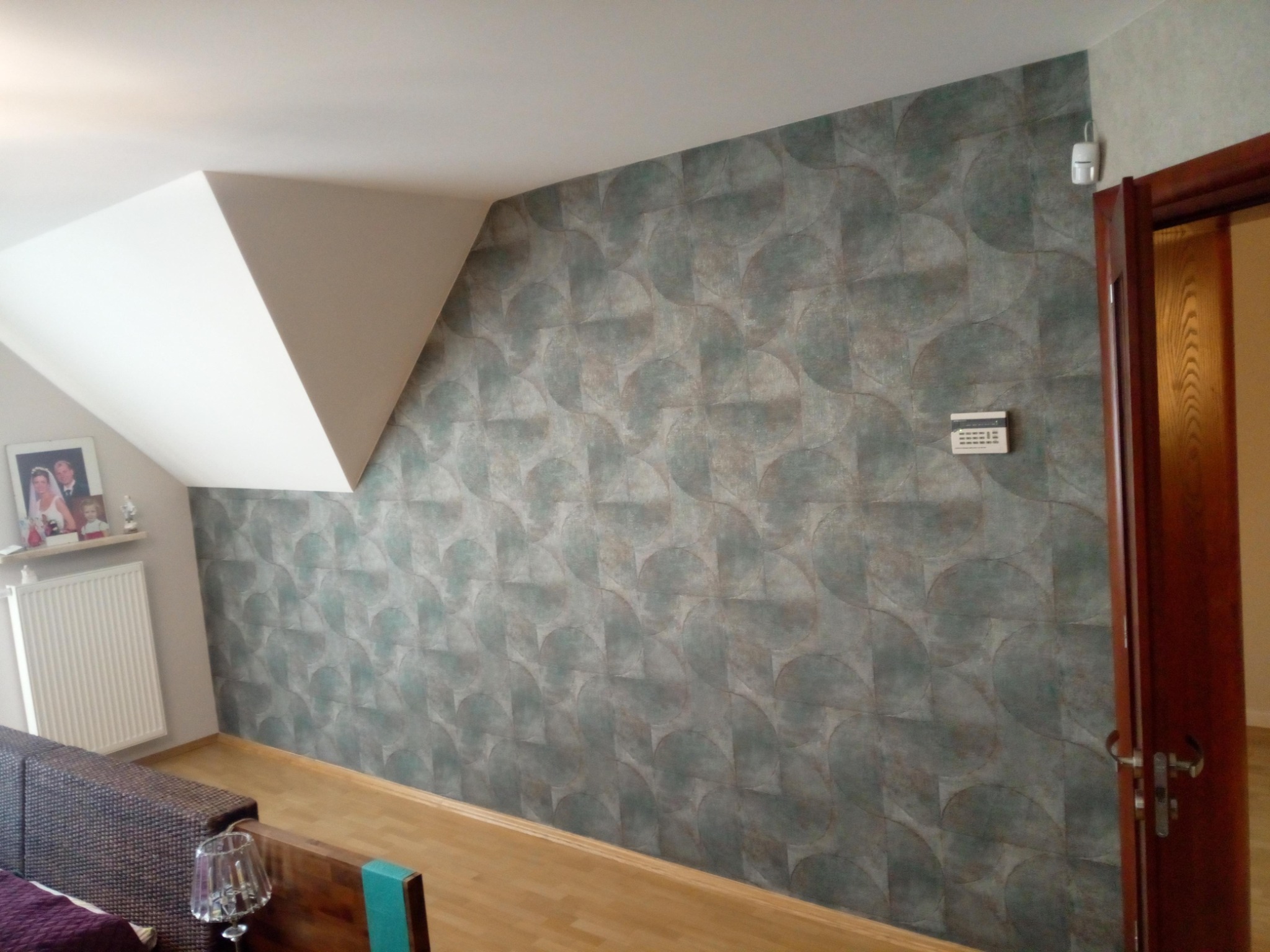 Pokój na poddaszu po położeniu tapety w odcieniach szarości z geometrycznym wzorem, widoczny narożnik ze skośnym sufitem i fragment kaloryfera.