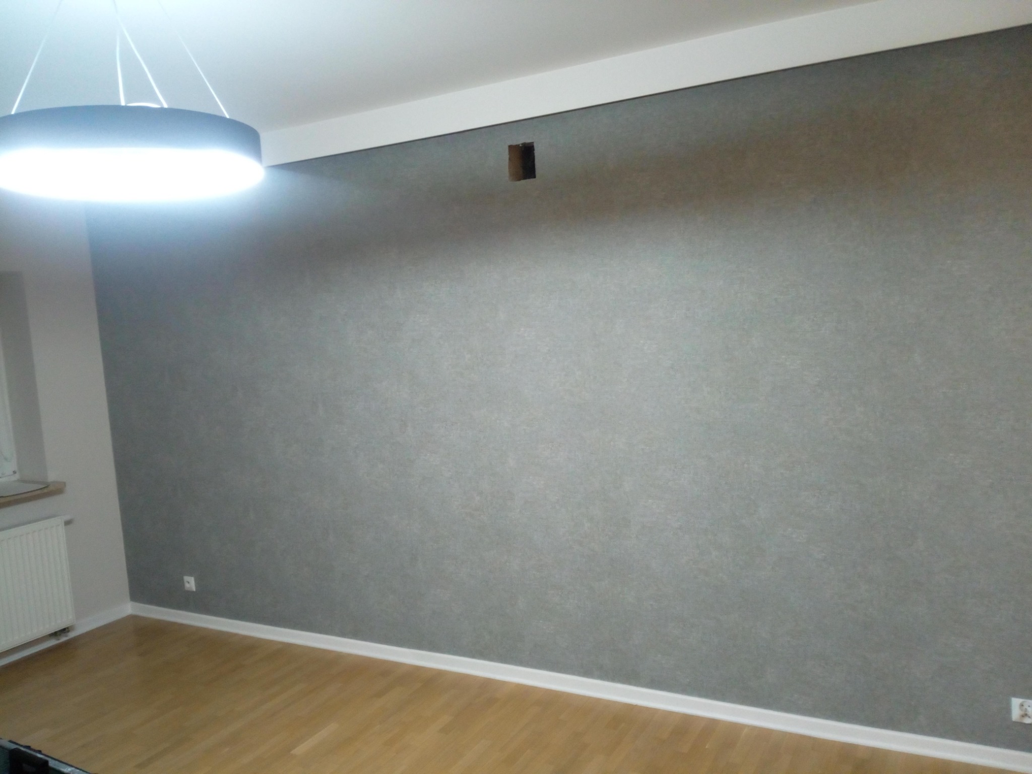 Ściana w pokoju po tapetowaniu, widoczna faktura tapety, otwór po gniazdku elektrycznym, drewniana podłoga i nowoczesna lampa sufitowa.