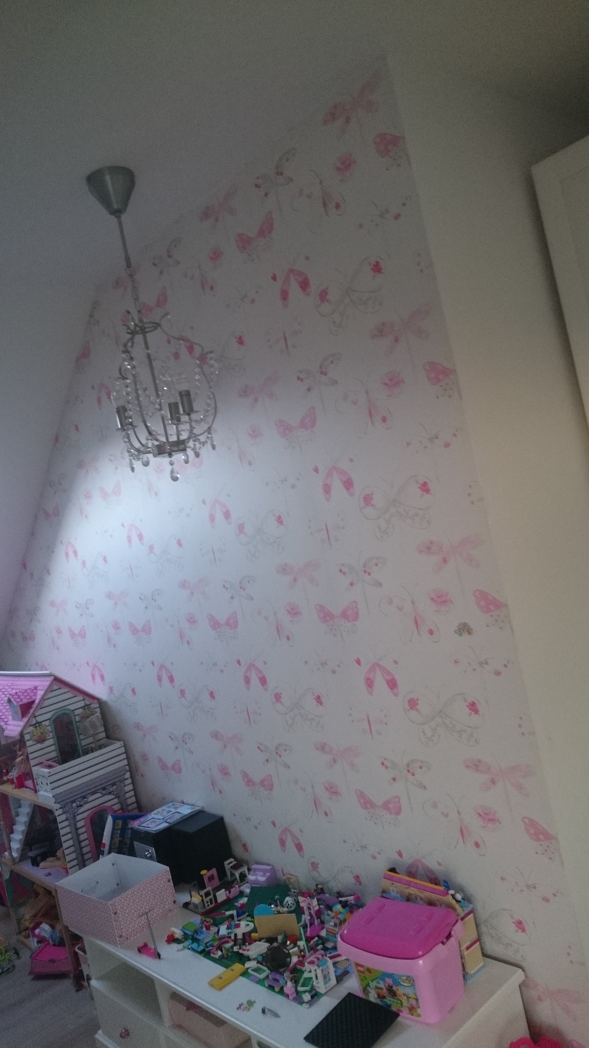 Pokój dziecięcy po tapetowaniu: biała ściana ze wzorem motyli, kryształowy żyrandol, zabawki na komodzie. Skośny sufit i fragment szafy w tle.
