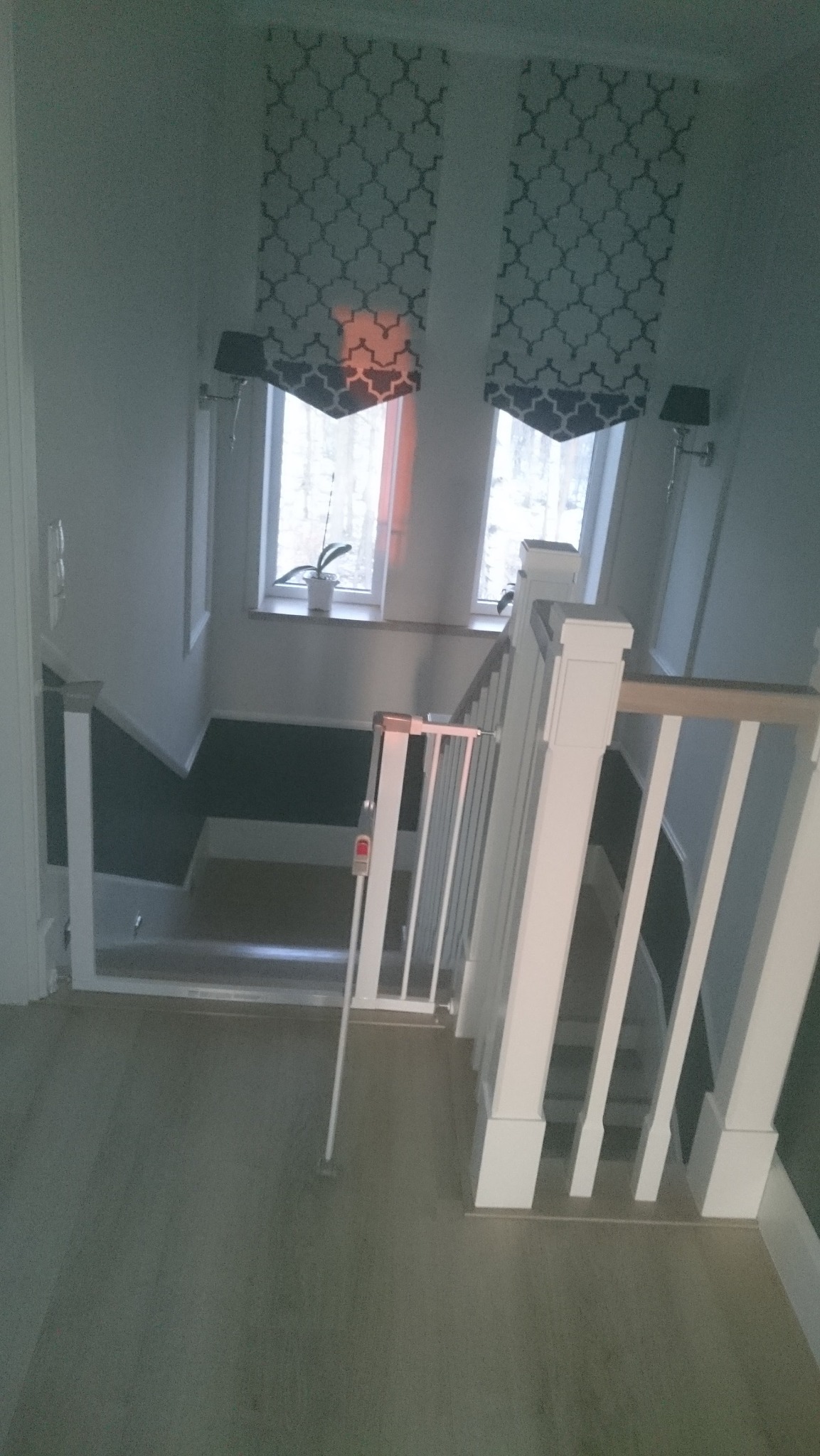 Klatka schodowa po odświeżeniu: białe balustrady, szare ściany z ciemniejszym pasem na dole, okna z dekoracyjnymi zasłonami i lampki ścienne.