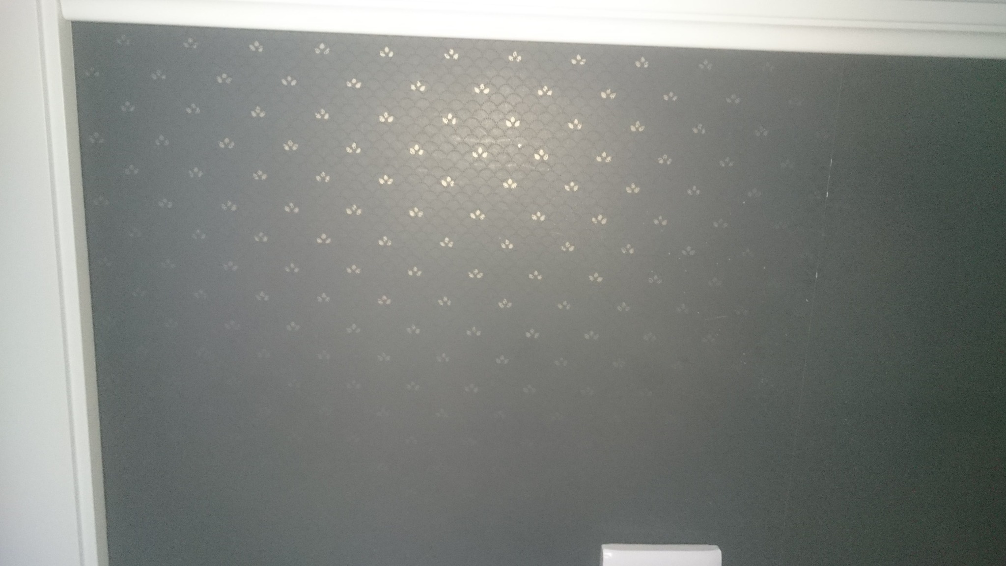 Szara ściana z delikatnym, srebrnym wzorem kwiatowym, wykończona białą listwą przypodłogową. Widoczny fragment gniazdka elektrycznego w dolnej części kadru.