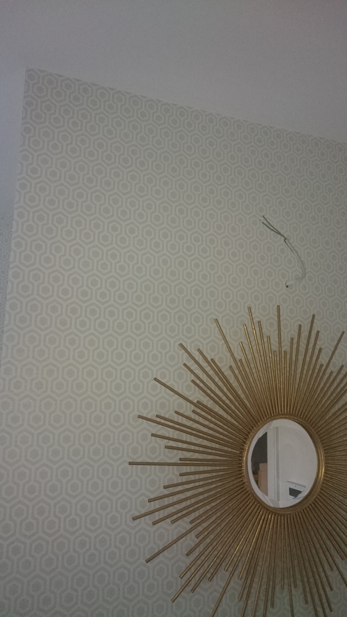Ściana z tapetą w geometryczny wzór, ozdobiona złotym lustrem w kształcie słońca. Widoczne wystające przewody elektryczne.
