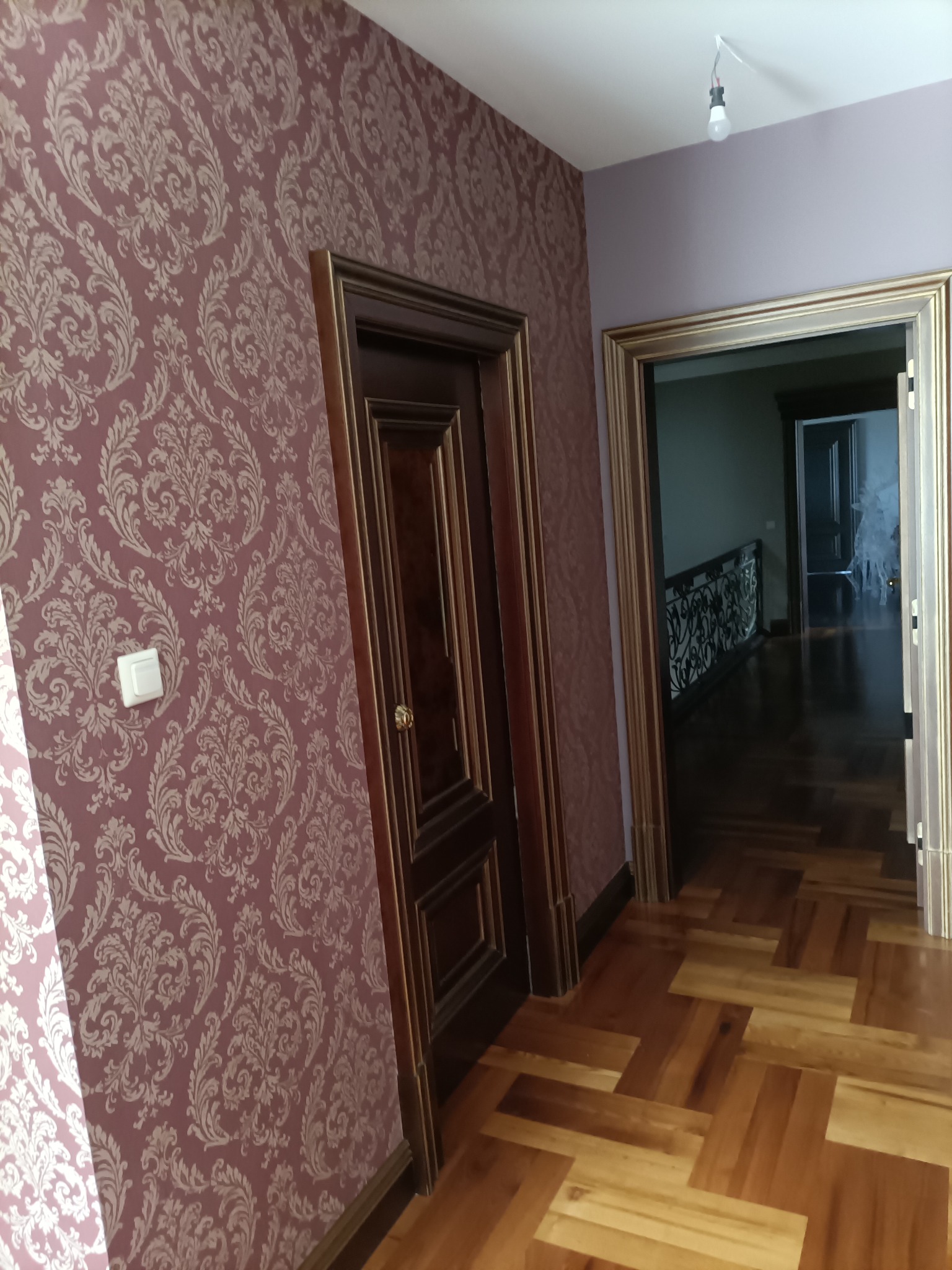 Korytarz po remoncie: wzorzysta tapeta w odcieniach różu i beżu, drewniane drzwi z ozdobnymi ramami, parkiet w jodełkę. Widok na dalszą część domu.
