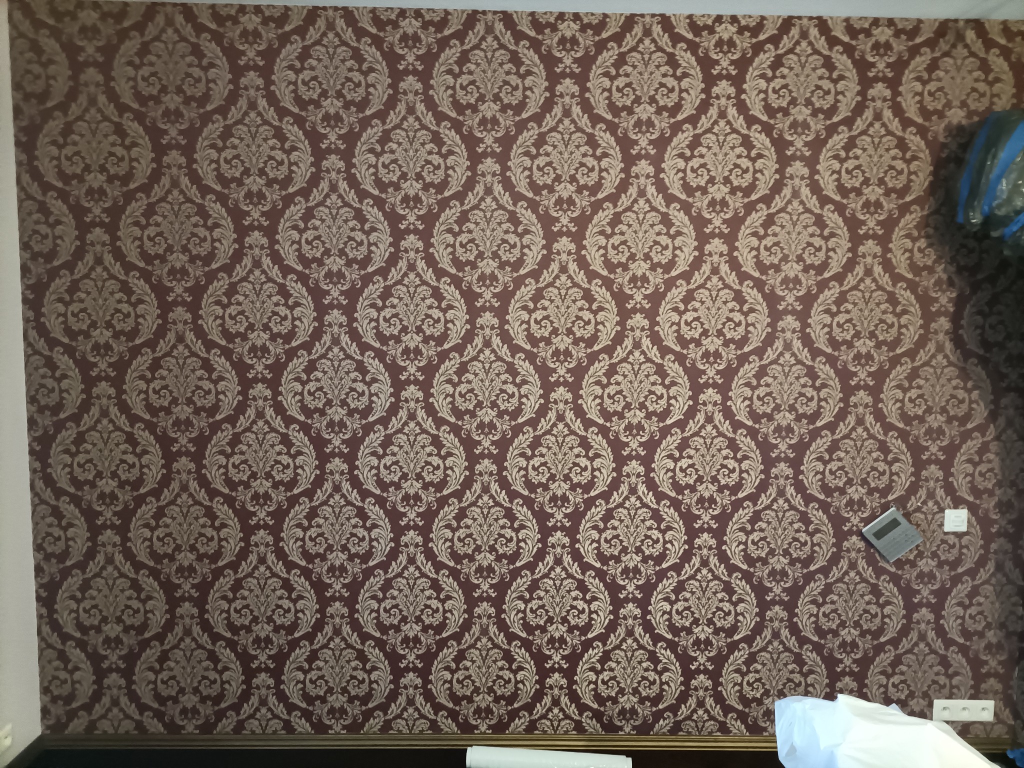 Ściana pokryta bordową tapetą w złote, symetryczne wzory. Widoczne gniazdka elektryczne i fragmenty przedmiotów przy ścianie. Dekoracyjne wykończenie wnętrza.