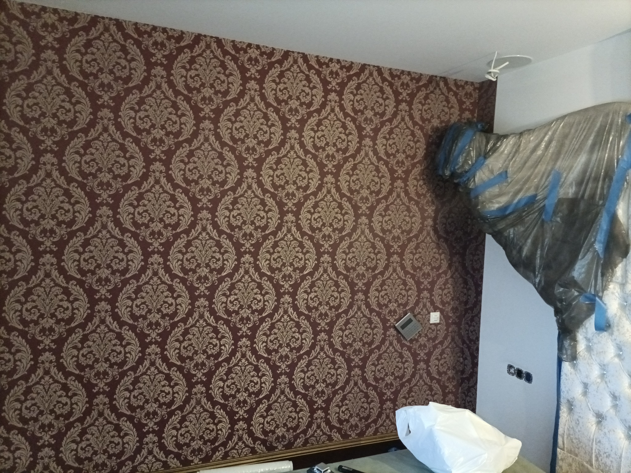 Ściana po tapetowaniu w brązowo-złote wzory. Część pomieszczenia zabezpieczona folią malarską. Widoczne gniazdka elektryczne i wystający element oświetlenia sufitowego.