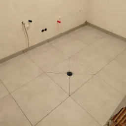 Jasne, kwadratowe płytki podłogowe ułożone w łazience z centralnie umieszczonym odpływem, widoczne wystające rurki instalacyjne na ścianie i fragment ściany w stanie surowym.