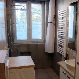 Jasna łazienka z szarymi płytkami, pralką pod drewnianym blatem, kabiną prysznicową typu walk-in z deszczownicą i białymi meblami łazienkowymi.